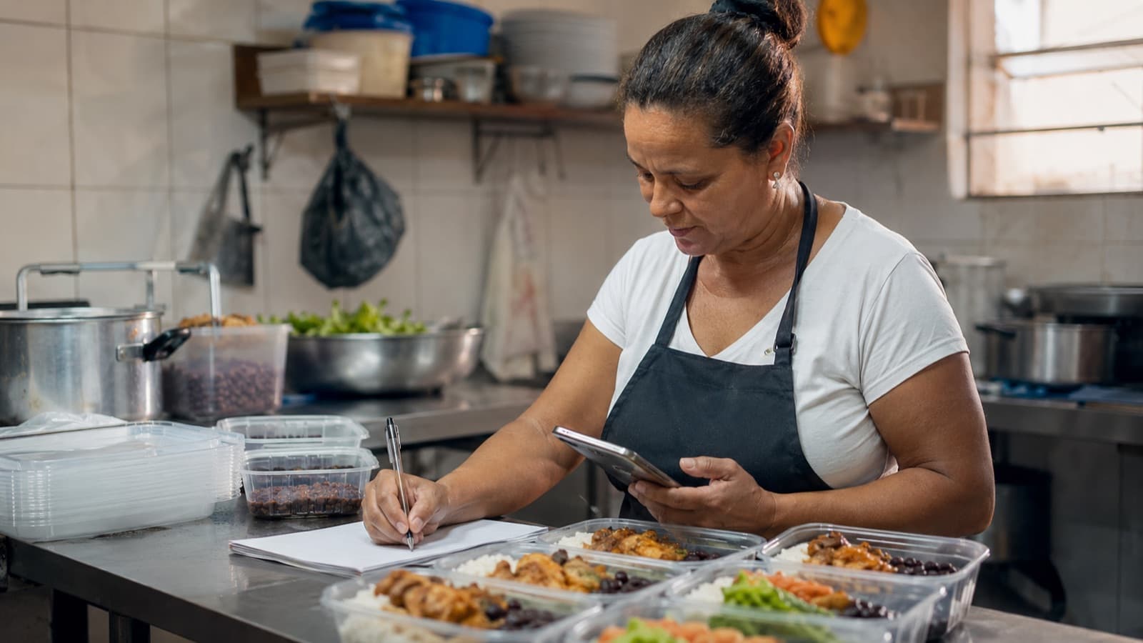 Dona de marmitaria anotando custos na cozinha, com marmitas montadas